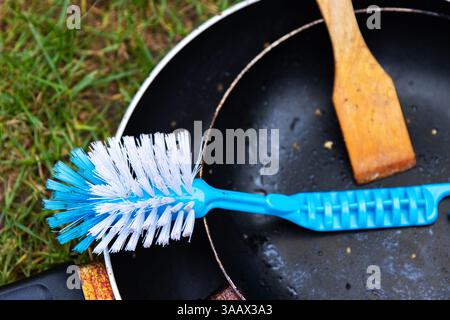 Draufsicht auf zwei schwarze Teflon-Pfannen, blaue Kunststoffbürste und Holzwäscher auf grünem Gras. Geschirrspülkonzept im Freien und Camping-Lifestyle. Co Stockfoto