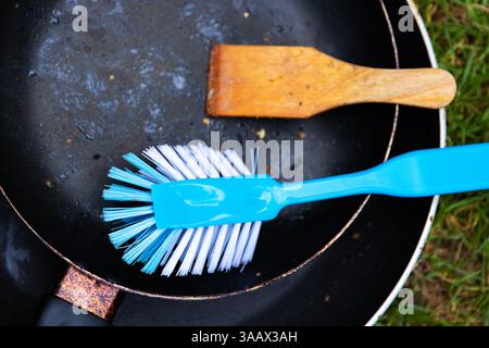 Draufsicht auf zwei schwarze Teflon-Pfannen, blaue Kunststoffbürste und Holzwäscher auf grünem Gras. Geschirrspülkonzept im Freien und Camping-Lifestyle. Co Stockfoto