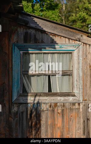 Nahaufnahme eines rustikalen Holzfensters mit weißen Vorhängen, eingerahmt von verwittertem Holz und beleuchtet von warmem Sonnenlicht. Das Bild erfasst ländliche CH Stockfoto