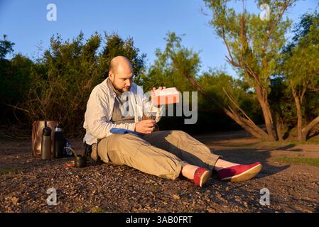 Der Reisende sitzt bei Sonnenaufgang am Ufer eines Baches und empfängt die ersten Sonnenstrahlen, während er sich darauf vorbereitet, Mate, ein typisches argentinisches Getränk, zu trinken. El Pal Stockfoto