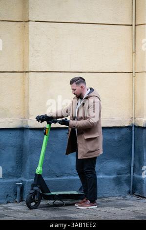 Ein junger Mann in einem stilvollen braunen Mantel steht neben einem leuchtend grünen Elektroroller und überprüft ihn, während er sich an eine strukturierte Stadtmauer lehnt Stockfoto