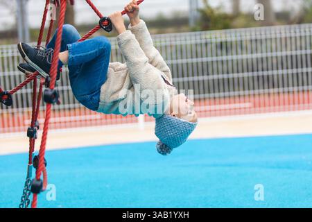 Ein verspieltes Kind in Strickmütze, Fleecejacke und Jeans hängt kopfüber an einer roten Kletterstruktur. Die hellblaue Oberfläche des Spielplatzes sorgt für einen zusätzlichen Stockfoto