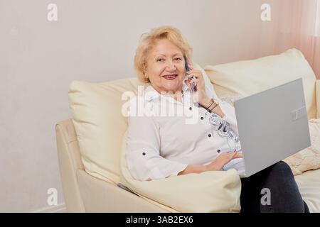 Seniorin, die bequem einen Laptop benutzt und mit ihrem Smartphone spricht, während sie zu Hause auf einem Sofa sitzt. Technologie, Kommunikation und Lebensstil der Senioren Stockfoto