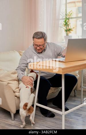 Senior Mann, der an einem Laptop an einem kleinen Tisch arbeitet, während er zu Hause seinen Jack Russell Terrier Hund streichelt. Arbeiten von zu Hause aus, Haustierbetreuung und Seniorenleben Stockfoto