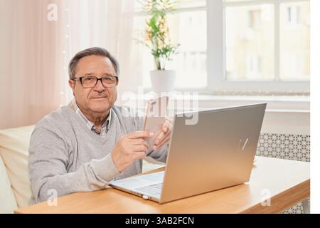 Gutaussehender, lächelnder Seniorenmann, der eine Brille mit Handy trägt, während er an seinem gemütlichen Arbeitsplatz mit Laptop zu Hause sitzt, pensionierter Mann, der mit freitag plaudert Stockfoto