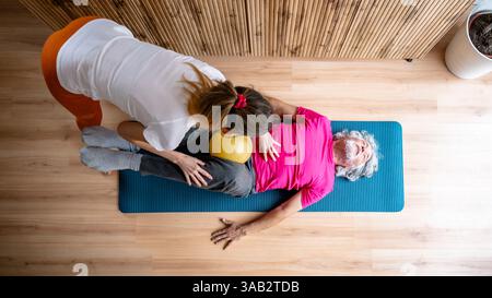 Eine Frau in orangefarbenen Leggings führt einen älteren Mann in einem rosafarbenen Hemd durch eine Kernübung mit einem gelben Ball, der in einer Fitnesssitzung auf einer blauen Matte liegt. Stockfoto