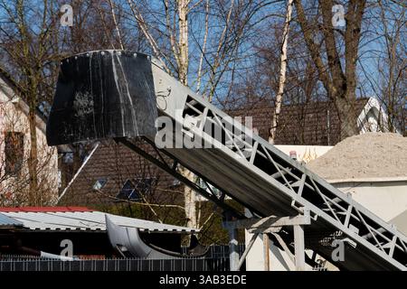 Ein über dem Boden angehobenes Förderband transportiert Sand von einer Pfanne zu einer Baustelle. Die Umgebung umfasst Wohngebäude und Bäume in Stockfoto