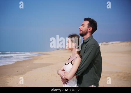 Romantisches Paar genießt eine friedliche Umarmung an einem breiten, leeren Strand, die Meeresbrise und die ruhige Aussicht auf die Küste genießen. Stockfoto