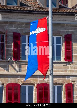 Nyon, Schweiz - 7. März 2025: Eine vertikale Flagge von Nyon mit blau-rotem Spaltmuster und weißem Fischemblem, die an einem Stab vor der Tür hängt Stockfoto
