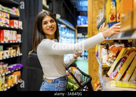 Junge Frau, die Lebensmittel kauft, eine Kiste aus dem Regal in einem Supermarkt, der auf Bio- und Naturprodukte spezialisiert ist, auswählt Stockfoto
