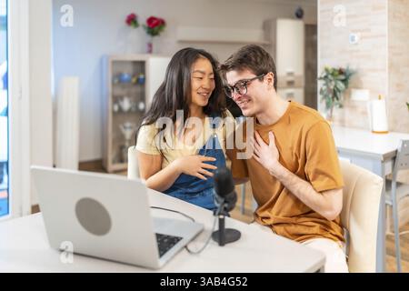 Junge, multiethnische Paare sitzen zu Hause zusammen und nehmen mit einem Laptop und Mikrofon ihren ansprechenden Audio-Podcast auf Stockfoto