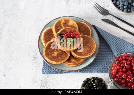 Hausgemachter Pfannkuchenstapel mit Heidelbeeren, Himbeeren und Erdbeeren auf einem weißen Teller mit einem blauen Tuch auf einer hellgrauen Tischplatte Stockfoto