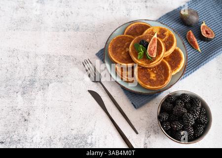 Hausgemachter Pfannkuchenstapel mit Heidelbeeren, Himbeeren und Erdbeeren auf einem weißen Teller mit einem blauen Tuch auf einer hellgrauen Tischplatte Stockfoto