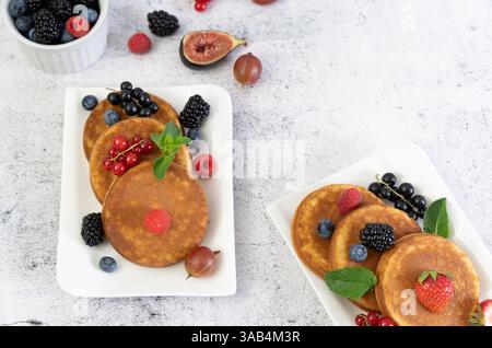 Klassische goldene Frühstückspancakes mit frischen Beeren auf zwei weißen quadratischen Tellern vor einer hellgrauen Tischplatte Stockfoto