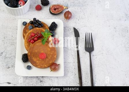 Süße und flauschige Pfannkuchen mit bunten Beeren, serviert auf einem modernen quadratischen Teller auf einer grauen Tischplatte Stockfoto