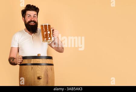 Ein bärtiger Mann trinkt köstliches Bier vom Fass im Pub oder in der Bar. Craft-Bier im Restaurant. Gutaussehender Mann mit einer Tasse Bier auf Holzfässern. Lächelnder Mann mit b Stockfoto