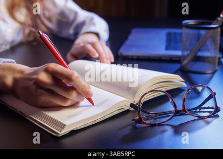 Eine Person, die mit einem roten Stift, einem Laptop und einem Stifthalter im Hintergrund in ein Notizbuch schreibt. Die Brille befindet sich auf dem Tisch neben dem Notebook. Stockfoto