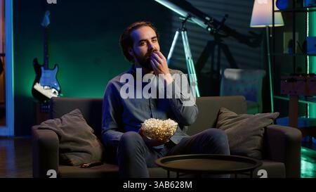 Mann, der den Fernseher mit der Fernbedienung anschaltet, die Freizeit zu Hause genießt und Popcorn genießt. Couch Potato Person in neonbeleuchtetem Apartment, isst salzigen Snack, unterhalten durch Fernsehen, Kamera A Stockfoto