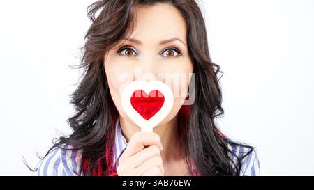 Eine junge Frau mit langen welligen brünetten Haaren trägt ein blau-weiß gestreiftes Hemd, das einen überraschenden Ausdruck zeigt, während sie ein herzförmiges Schild in f hält Stockfoto