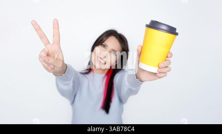 Lächelnde Frau, die eine gelbe Mockup-Kaffeetasse hochhält, mit der Hand eines Stückschilds vor einem weißen Hintergrund. Stockfoto