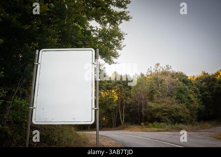 Leeres weißes Straßenschild neben einer ländlichen kroatischen Straße, umgeben von dichtem Wald, bietet ausreichend Platz für benutzerdefinierte Richtungs- oder Informationstexte. Stockfoto