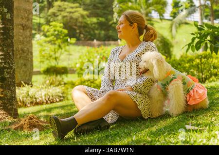 Reife Frau, die auf dem Gras in einem lebhaften Park sitzt und die Sonne genießt, während sie die Gesellschaft ihres kleinen weißen Pudels genießt, der in einem Colo gekleidet ist Stockfoto