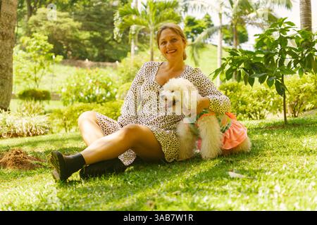 Eine Frau mittleren Alters genießt einen friedlichen Moment in einem lebhaften Park, sitzt mit ihrem kleinen, weißen Hund in einem bunten Kleid auf dem Gras, umgeben von l Stockfoto