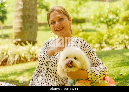 Glückliche Frau, die ihren süßen weißen Bigon-Friseurhund umschließt, während sie einen sonnigen Tag in einem Park genießt und die starke Bindung zwischen Haustier und Besitzer demonstriert Stockfoto