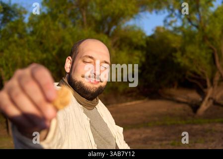Der Mann lächelt und bietet der Kamera draußen einen Keks an. Komposition mit selektivem Fokus auf das Gesicht. Stockfoto