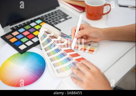 Nahaufnahme einer Hand, die mit einem Stift auf eine Farbe auf der Farbpalette zeigt. Auswählen oder Auswählen einer Farbe. Innenarchitekt oder Grafikdesigner, der im Büro arbeitet Stockfoto