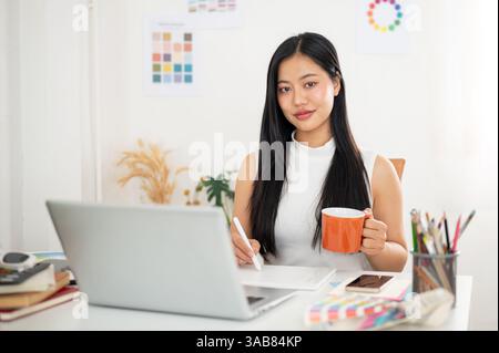 Eine asiatische Frau hält einen Stift, zeichnet oder schreibt auf Tablets in der einen Hand und hält einen Becher in der anderen Hand. Innenarchitekt oder Grafikdesigner Stockfoto