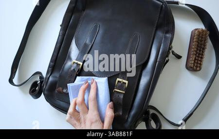 Die Hand einer Frau, die einen verrückten Pferdeleder-Rucksack reinigt. Schwarzes Leder. Goldene Hardware. Wachs und eine Reinigungsbürste in der Nähe. Die weiße Hand einer Frau wischt die Tasche mit einem Schwamm ab. Weißer Hintergrund. Foto. Stockfoto
