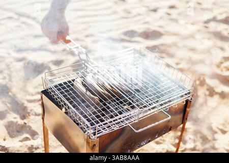 Nahaufnahme eines Mannes, der einen Handgrill mit Bratwürsten hält. Grill am Sandstrand. Ein Mann brät Würstchen auf offenem Feuer. Picknick auf t Stockfoto