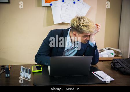 Ein junger Mann mit blonden Haaren und blauen Augen sitzt an einem Schreibtisch und sieht konzentriert aus, während er an seinem Laptop arbeitet. Papiere und Geräte umgeben ihn in dem hellen, modernen Büro. Stockfoto