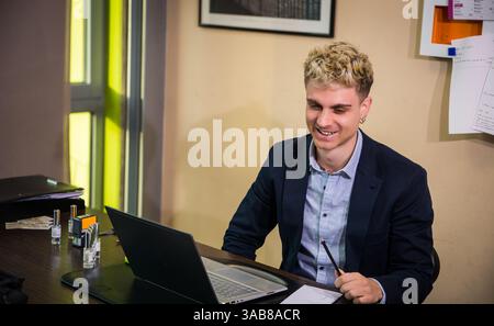 Ein junger Mann mit attraktiven Funktionen sitzt in einem eleganten Büro und konzentriert sich auf seinen Laptop-Bildschirm. Er trägt einen marineblauen Blazer und lächelt, während er einen Stift hält, umgeben von Papieren und Bürobedarf. Stockfoto