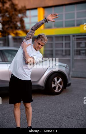 Ein hübscher junger Mann mit blonden Haaren und blauen Augen streckt seine Arme, während er während des Sonnenuntergangs neben einem silbernen Auto steht. Sein sportlicher Auftritt unterstreicht einen aktiven Lebensstil. Stockfoto
