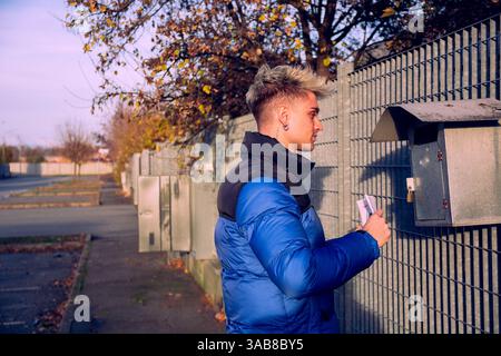 Ein junger, attraktiver Mann mit blonden Haaren und blauen Augen steht neben einem Briefkasten. Er hält Briefe und freut sich, seine Post an einem klaren, sonnigen Tag in einer friedlichen Gegend zu finden. Stockfoto