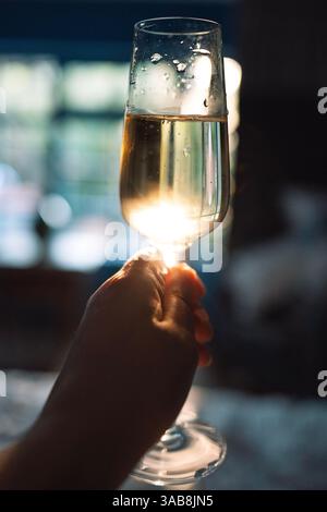 Nahaufnahme einer weiblichen Hand, die ein elegantes Glas Champagner mit Toast hält. Eine Frau hält eine Tasse Sekt in den Strahlen der untergehenden Sonne A Stockfoto