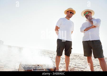 Zwei junge Männer in lässiger Sommerkleidung stehen neben einem Grill am Meer. Freunde lachen und grillen Fleisch am Sandstrand. Lächelnde Männer i Stockfoto