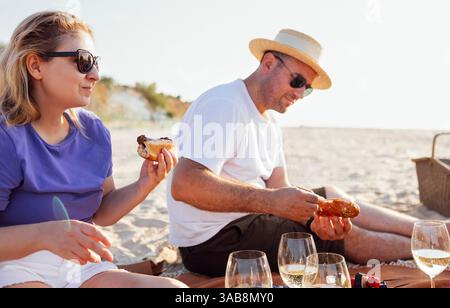 Ein lächelnder Mann und eine Frau sitzen am Meer und machen ein Picknick. Ein verheiratetes Paar isst und hat Spaß mit Freunden an einem Sandstrand. Menschen Stockfoto