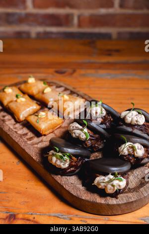 Elegante Portion von schwarzen Kraken bao-Brötchen mit cremiger Sauce und Mikrogrün, gepaart mit goldbraunen Vorspeisen, kunstvoll auf einem rustikalen Ambiente angeordnet Stockfoto