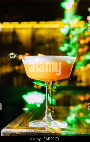 Ein lebhafter Pfirsichcocktail mit einer Schaumplatte, elegant in einem Coupé-Glas serviert. Der Cocktail sitzt auf einer reflektierenden Glasbar, die von warmem Gold beleuchtet wird Stockfoto