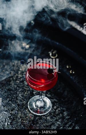 Ein tiefroter Gin-Cocktail mit Beeren, blumigen Noten und Sektspritzern, elegant garniert mit einer dunklen Kirsche. Gegen eine rauchige Rückseite Stockfoto