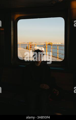 Ein Mann sitzt ruhig in einer Fähre und blickt auf den Sonnenuntergang über dem Wasser in New York. Das warme Glühen sorgt für eine ruhige Atmosphäre, die die Fortsetzung unterstreicht Stockfoto
