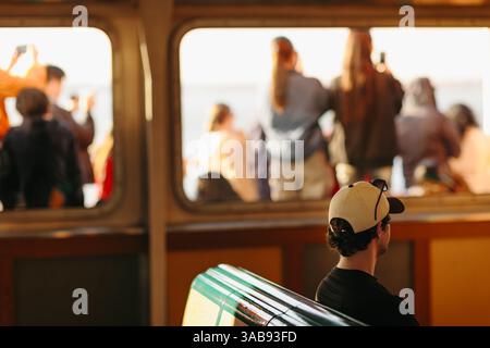 Ein Mann sitzt in einer Fähre mit einer Kappe, während andere Passagiere am Fenster stehen und den Sonnenuntergang mit Smartphones in New York einfangen. Das Atmo Stockfoto