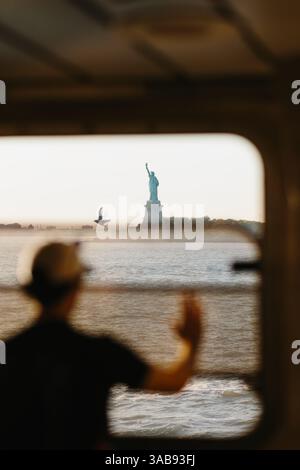 Verschwommene Silhouette eines Mannes, der in einer Fähre in New York winkt. Die Freiheitsstatue und eine fliegende Möwe sind draußen zu sehen. Die Szene fängt eine Mutter ein Stockfoto