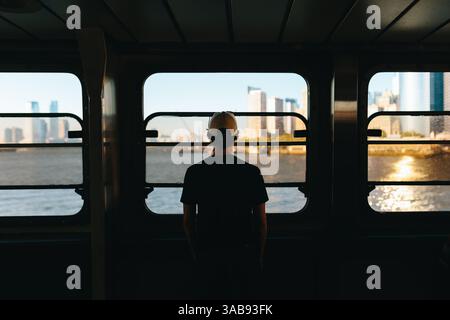 Ein Mann mit Mütze steht auf einer Fähre und blickt auf eine glitzernde Skyline der Stadt über dem Wasser in New York. Die Szene erfasst Introspektion und urba Stockfoto