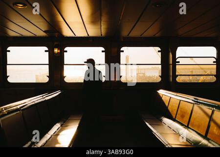 Ein Mann in Silhouette steht in einer Fähre und blickt auf eine ruhige, sonnendurchflutete Hafenszene in New York. Das warme, goldene Sonnenlicht erzeugt einen atmosphärischen Glo Stockfoto