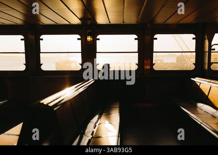 Ein einsamer Reisender sitzt auf einer Fähre, umgeben von einem warmen Sonnenuntergang in New York. Das Licht filtert durch die Fenster und erzeugt ein beruhigendes Licht in t Stockfoto