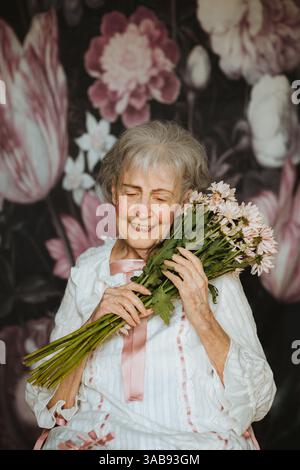 Eine ältere Frau mit einem fröhlichen Ausdruck hält einen Blumenstrauß, umgeben von einem floralen Hintergrund. Ihr Glück strahlt in diesem herzerwärmenden stu aus Stockfoto
